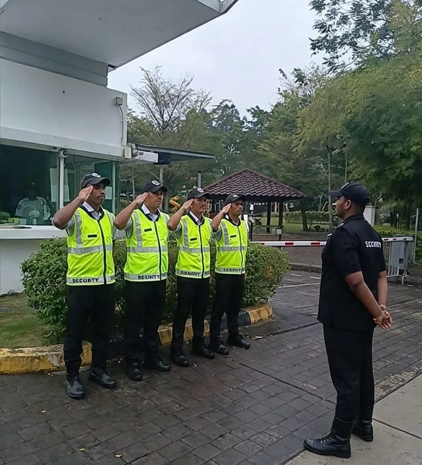 Four guards in hi-visibility vests saluting a supervisor beside a guardhouse